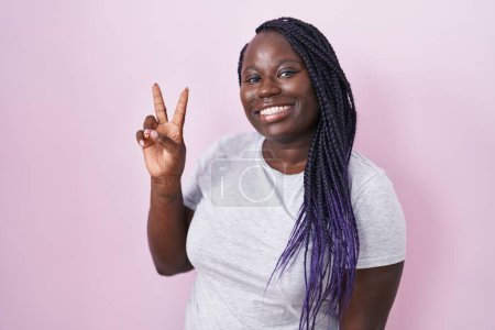 Foto de Mujer africana joven de pie sobre fondo rosa sonriendo mirando a la cámara mostrando los dedos haciendo signo de victoria. número dos. - Imagen libre de derechos