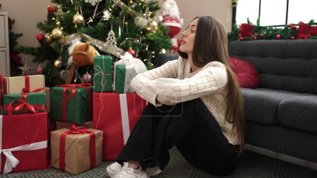 Téléchargez les photos : Jeune belle femme hispanique assise près de l'arbre de Noël avec une expression sérieuse à la maison - en image libre de droit