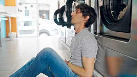 Téléchargez les photos : Jeune homme hispanique attendant une machine à laver avec un visage sérieux à la laverie - en image libre de droit