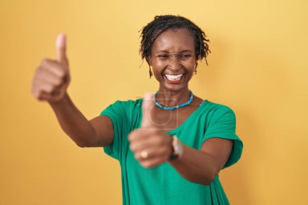 Téléchargez les photos : Femme africaine avec dreadlocks debout sur fond jaune approuvant faire un geste positif avec la main, pouces levés souriant et heureux pour le succès. geste gagnant. - en image libre de droit