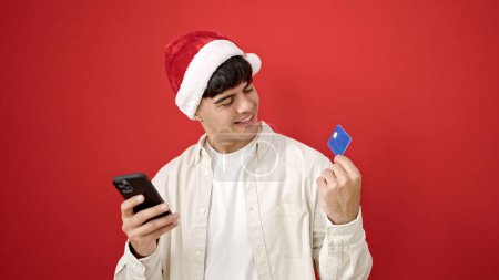 Foto de Joven hombre hispano de compras con teléfono inteligente y tarjeta de crédito con sombrero de Navidad sobre fondo rojo aislado - Imagen libre de derechos