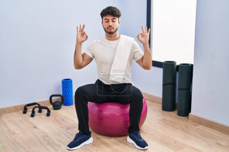 Téléchargez les photos : Homme hispanique avec barbe assis sur des boules de pilate à la salle de yoga se détendre et sourire les yeux fermés en faisant geste de méditation avec les doigts. concept de yoga. - en image libre de droit