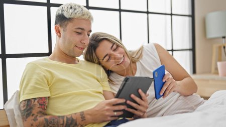 Photo for Beautiful couple using smartphone and touchpad sitting on bed at bedroom - Royalty Free Image