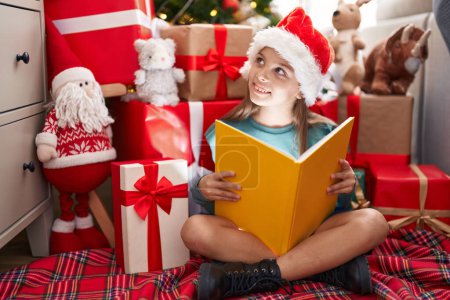 Photo for Adorable hispanic girl reading book sitting on floor by christmas tree at home - Royalty Free Image