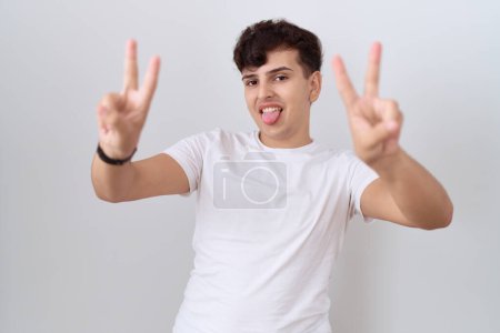 Foto de Joven hombre no binario con camiseta blanca casual sonriendo con la lengua hacia fuera mostrando los dedos de ambas manos haciendo signo de victoria. número dos. - Imagen libre de derechos