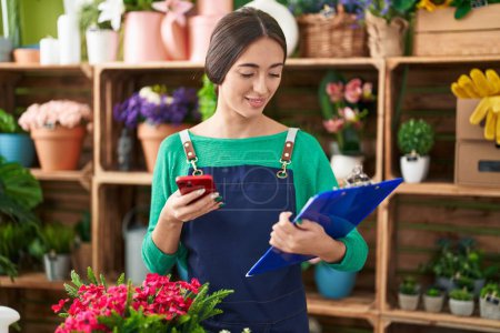 Photo for Young beautiful hispanic woman florist using smartphone reading document at flower shop - Royalty Free Image