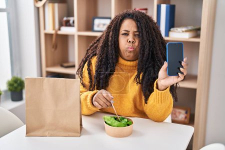Téléchargez les photos : Plus taille hispanique femme manger emporter salade tenant smartphone joues gonflées avec drôle de visage. bouche gonflée d'air, attrapant l'air. - en image libre de droit