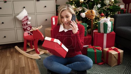 Photo for Young blonde woman talking on smartphone holding christmas gift at home - Royalty Free Image