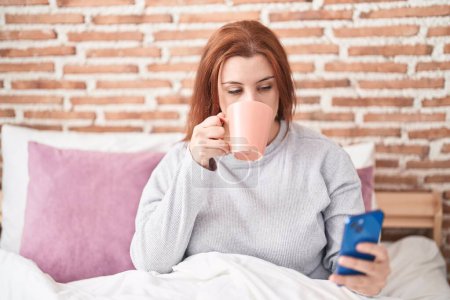 Téléchargez les photos : Young beautiful plus size woman using smartphone drinking coffee at bedroom - en image libre de droit