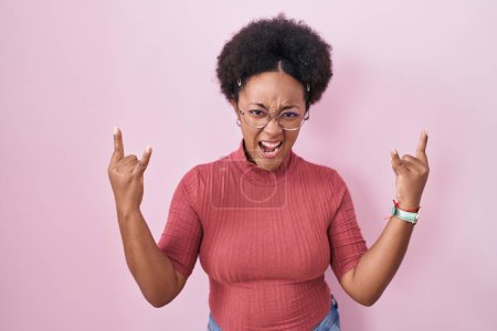 Photo for Beautiful african woman with curly hair standing over pink background shouting with crazy expression doing rock symbol with hands up. music star. heavy music concept. - Royalty Free Image