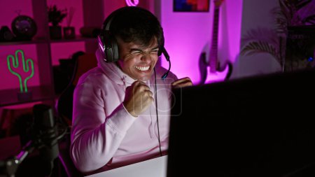 Téléchargez les photos : Jeune hispanique attrayant gagnant au streaming, célébrant la victoire du jeu vidéo avec un grand sourire dans la salle de jeu sombre - en image libre de droit