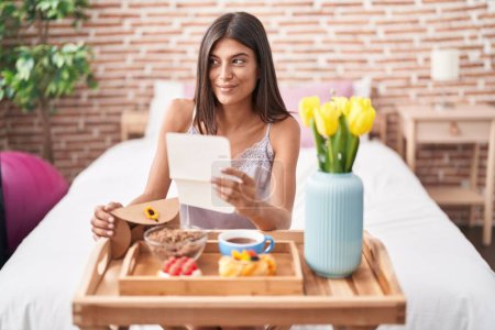 Foto de Morena mujer joven desayunando en la cama leyendo una carta sonriendo mirando a un lado y mirando hacia otro pensando. - Imagen libre de derechos