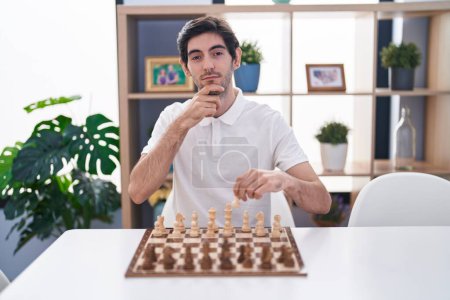 Foto de Joven hombre hispano jugando ajedrez sentado en la mesa cara seria pensando en la pregunta con la mano en la barbilla, pensativo acerca de la idea confusa - Imagen libre de derechos