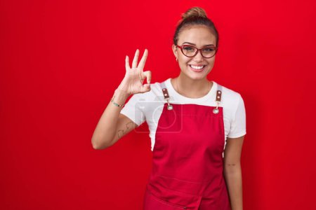 Photo for Young hispanic woman wearing waitress apron over red background smiling positive doing ok sign with hand and fingers. successful expression. - Royalty Free Image