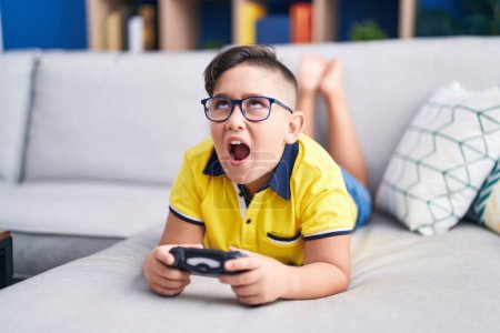 Photo for Young hispanic kid playing video game holding controller on the sofa angry and mad screaming frustrated and furious, shouting with anger looking up. - Royalty Free Image