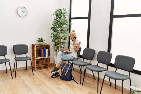 Téléchargez les photos : Jeune femme blonde parlant sur smartphone écrivant sur un document assis sur une chaise dans la salle d'attente - en image libre de droit