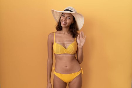 Foto de Mujer hispana joven con bikini y sombrero de verano renunciando a decir hola feliz y sonriente, gesto de bienvenida amistoso - Imagen libre de derechos