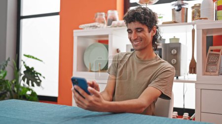 Photo for Young hispanic man using smartphone sitting on table at dinning room - Royalty Free Image