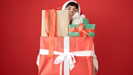 Téléchargez les photos : Jeune homme hispanique portant un chapeau de Noël tenant des cadeaux sur fond rouge isolé - en image libre de droit