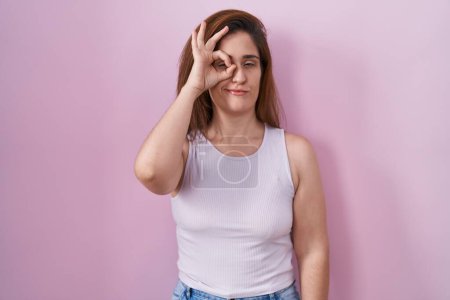 Photo for Brunette woman standing over pink background doing ok gesture with hand smiling, eye looking through fingers with happy face. - Royalty Free Image