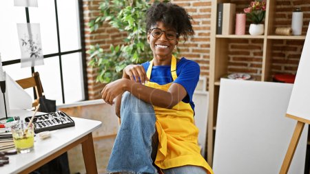 Téléchargez les photos : Artiste afro-américaine souriante confiante assise sur une chaise au studio d'art - en image libre de droit