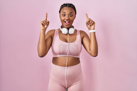 Foto de Mujer afroamericana con trenzas con ropa deportiva y auriculares sonriendo asombrada y sorprendida y señalando con los dedos y los brazos levantados. - Imagen libre de derechos