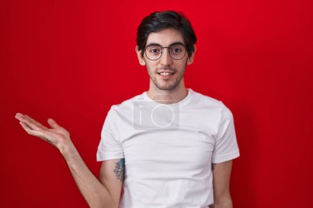 Téléchargez les photos : Jeune homme hispanique debout sur fond rouge souriant gai présentation et pointant avec la paume de la main en regardant la caméra. - en image libre de droit