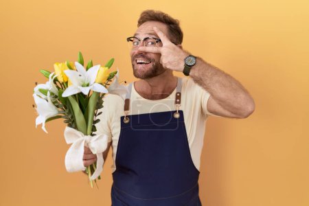 Téléchargez les photos : Homme du Moyen Age avec magasin de fleuriste barbe tenant des fleurs faisant symbole de paix avec les doigts sur le visage, souriant joyeux montrant la victoire - en image libre de droit