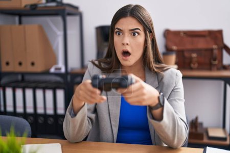 Photo for Hispanic woman working at the office playing video games in shock face, looking skeptical and sarcastic, surprised with open mouth - Royalty Free Image