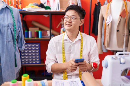 Téléchargez les photos : Jeune chinois tailleur souriant confiant en utilisant smartphone à l'atelier - en image libre de droit