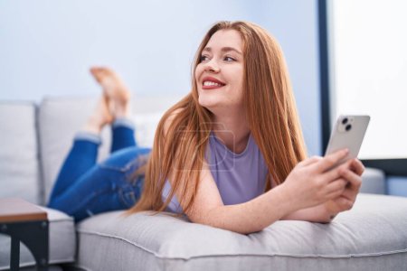 Téléchargez les photos : Jeune rousse femme en utilisant smartphone couché sur le canapé à la maison - en image libre de droit