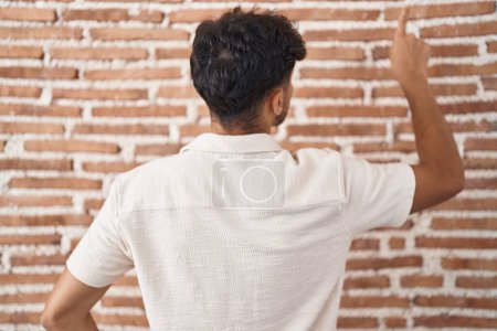 Téléchargez les photos : Homme arabe avec barbe debout sur fond de mur de briques posant vers l'arrière pointant vers l'avant avec la main du doigt - en image libre de droit