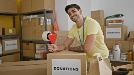 Téléchargez les photos : Jeune homme hispanique volontaire souriant confiant appuyé sur les dons boîte en carton tenant bande d'emballage au centre de bienfaisance - en image libre de droit