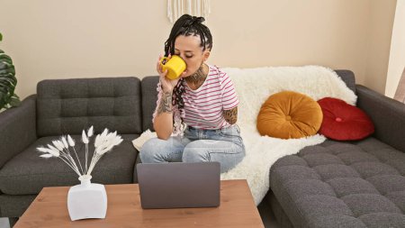 Foto de Mujer amputada hispana, seriamente enfocada, utilizando la computadora portátil en casa; sentada en el sofá, bebiendo café, tatuajes trazando su lado sin brazos. - Imagen libre de derechos