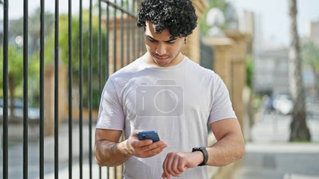 Téléchargez les photos : Jeune homme latin utilisant smartphone et smartwatch à la rue - en image libre de droit