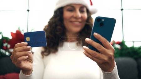 Foto de Mujer hispana de mediana edad usando teléfono inteligente y tarjeta de crédito sentado en el sofá por la decoración de Navidad en casa - Imagen libre de derechos