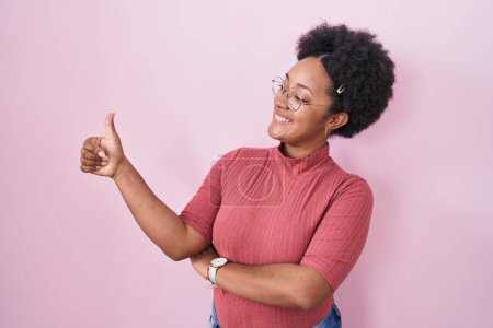 Téléchargez les photos : Belle femme africaine avec les cheveux bouclés debout sur fond rose regardant fier, souriant faire les pouces geste vers le haut sur le côté - en image libre de droit