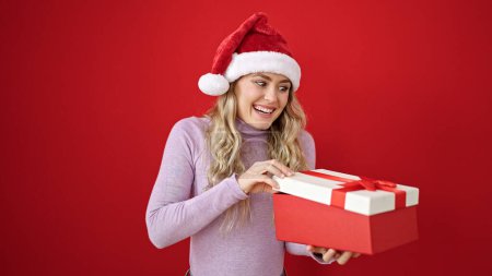 Téléchargez les photos : Jeune femme blonde portant un chapeau de Noël déballer cadeau sur fond rouge isolé - en image libre de droit