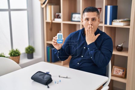 Téléchargez les photos : Jeune homme hispanique tenant dispositif glucomètre couvrant la bouche avec la main, choqué et peur de l'erreur. expression surprise - en image libre de droit