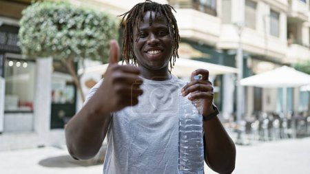Téléchargez les photos : Afro-Américain tenant une bouteille d'eau faisant un geste de pouce sur la terrasse du café - en image libre de droit