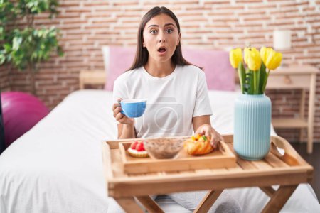 Téléchargez les photos : Jeune femme hispanique manger des pâtisseries pour le petit déjeuner assis sur le lit effrayé et choqué par la surprise et l'expression étonnante, la peur et le visage excité. - en image libre de droit