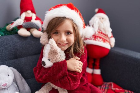 Photo for Adorable hispanic girl hugging teddy bear sitting on sofa by christmas decor at home - Royalty Free Image