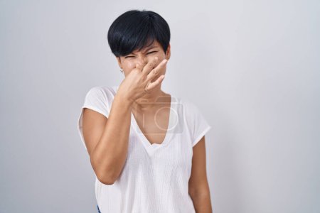 Foto de Mujer asiática joven con el pelo corto de pie sobre el fondo aislado oliendo algo apestoso y asqueroso, olor intolerable, conteniendo la respiración con los dedos en la nariz. mal olor - Imagen libre de derechos
