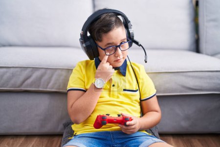 Téléchargez les photos : Jeune enfant hispanique jouant contrôleur de tenue de jeu vidéo portant des écouteurs pointant vers l'œil vous regardant geste, expression suspecte - en image libre de droit