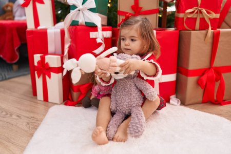 Foto de Adorable niño rubio sosteniendo oso de peluche sentado en el suelo por regalos de Navidad en casa - Imagen libre de derechos