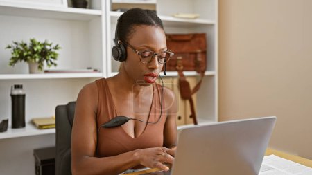 Téléchargez les photos : Belle femme afro-américaine, travailleuse d'affaires concentrée, aide à la parole dans un casque tout en se concentrant sur un ordinateur portable, dans un intérieur de bureau détendu - en image libre de droit