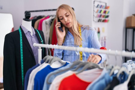 Téléchargez les photos : Jeune femme blonde tailleur tenant des vêtements sur rack parler sur smartphone au magasin de tailleur - en image libre de droit