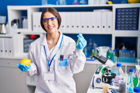Foto de Joven hermosa mujer hispana científica sosteniendo limón y tubo de ensayo en el laboratorio - Imagen libre de derechos