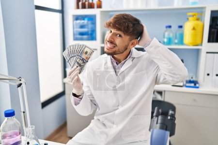 Foto de Hombre árabe con barba trabajando en laboratorio científico sosteniendo dinero estresado y frustrado con la mano en la cabeza, rostro sorprendido y enojado - Imagen libre de derechos