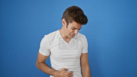 Téléchargez les photos : Jeune homme hispanique souffrant de maux d'estomac debout sur fond bleu isolé - en image libre de droit